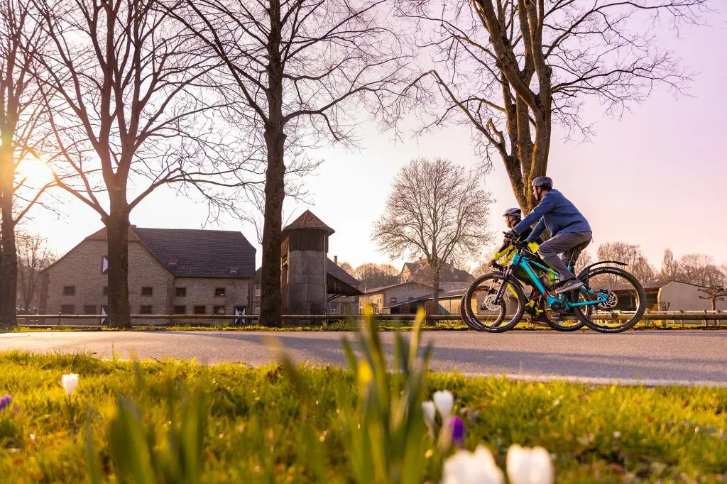 Zwei Radfahrer auf einer Straße mit einem Dorf im Hintergrund.