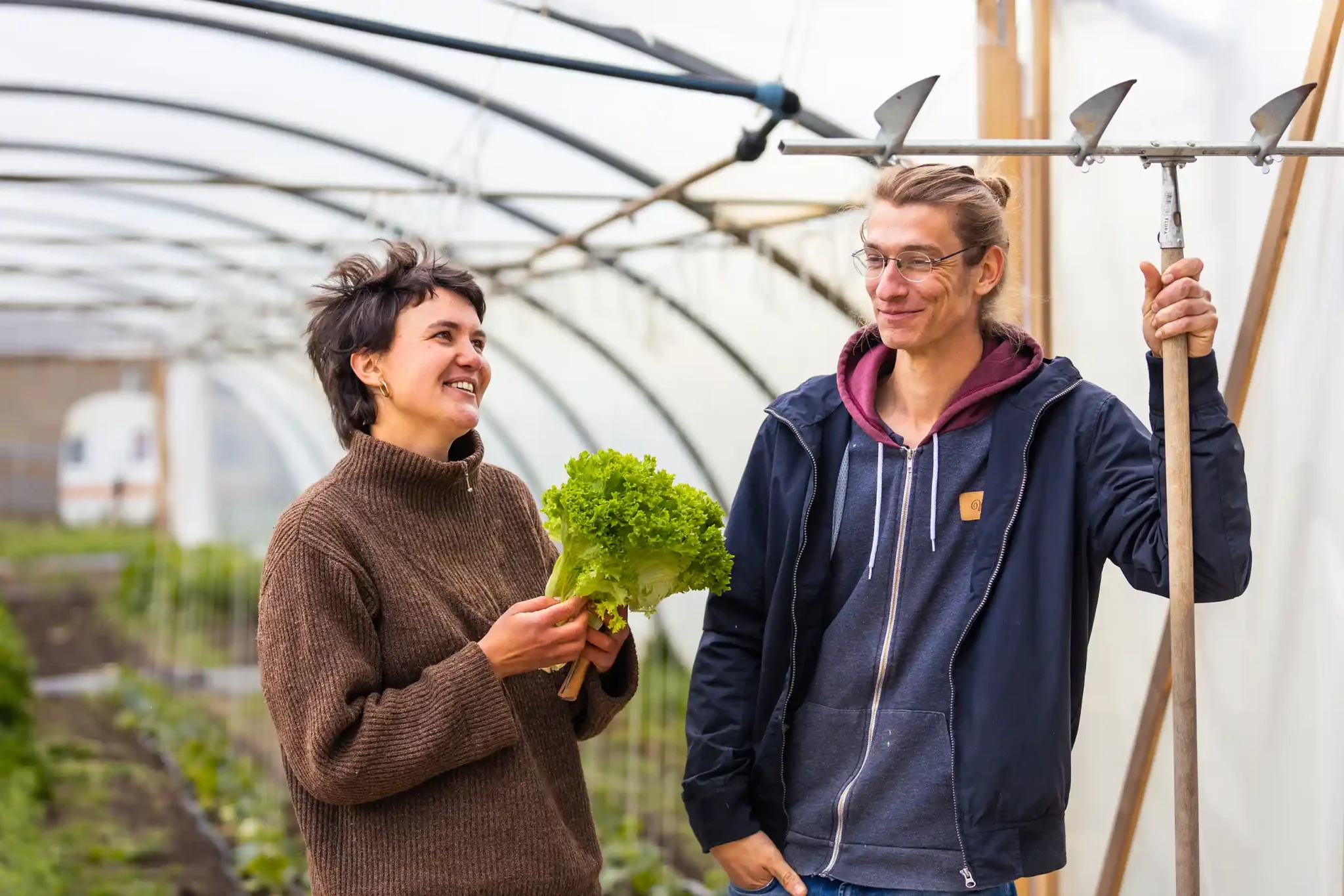 Malte und eine Frau stehen zusammen im Gewächshaus, Malte hält Gartenwerkzeug, die Frau einen frischen Salat, beide lächeln.