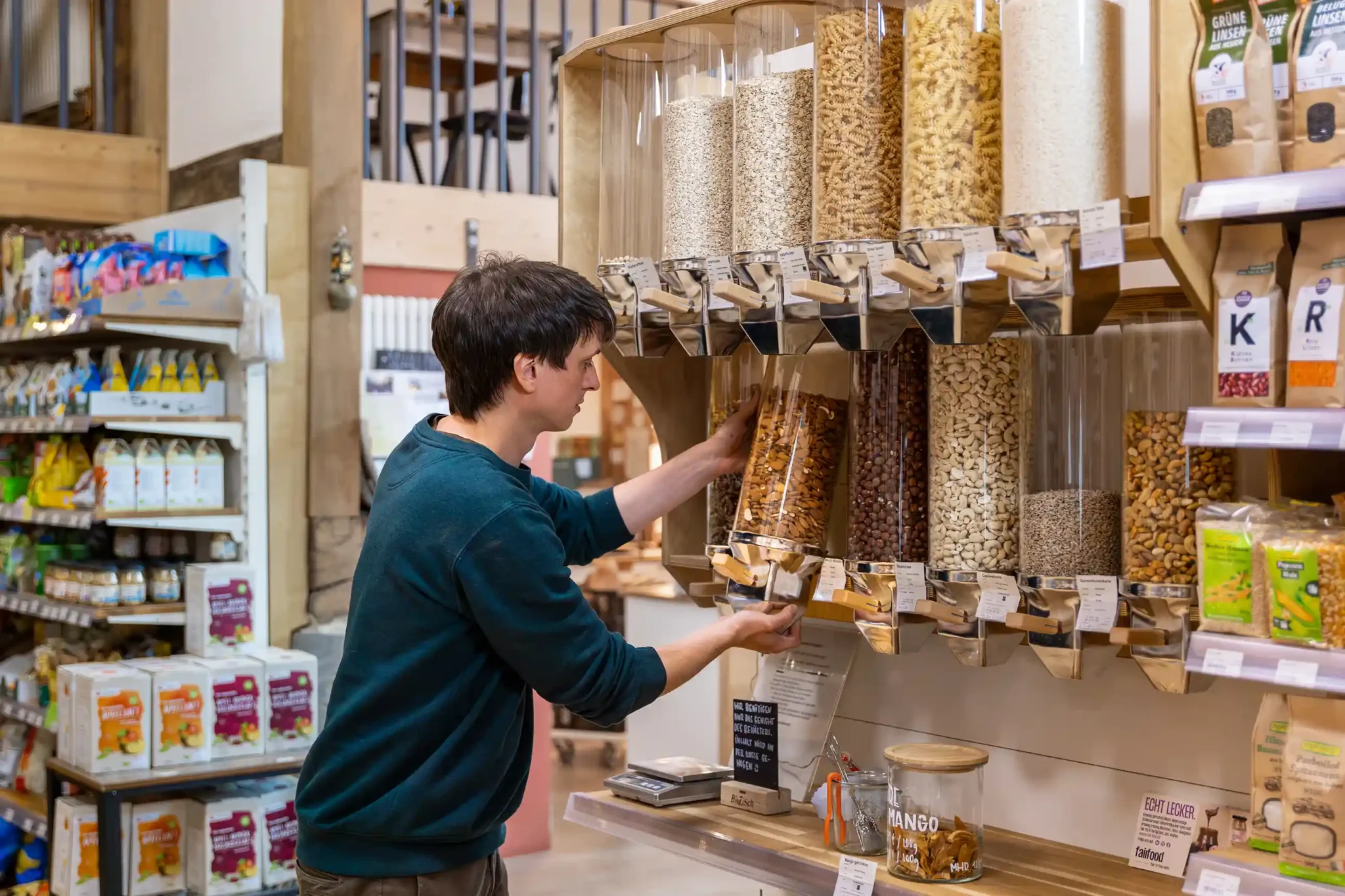 Ein Mann füllt Nüsse in einem Glasbehälter im Hofladen auf, umgeben von Holzregalen mit nachhaltigen Lebensmitteln.