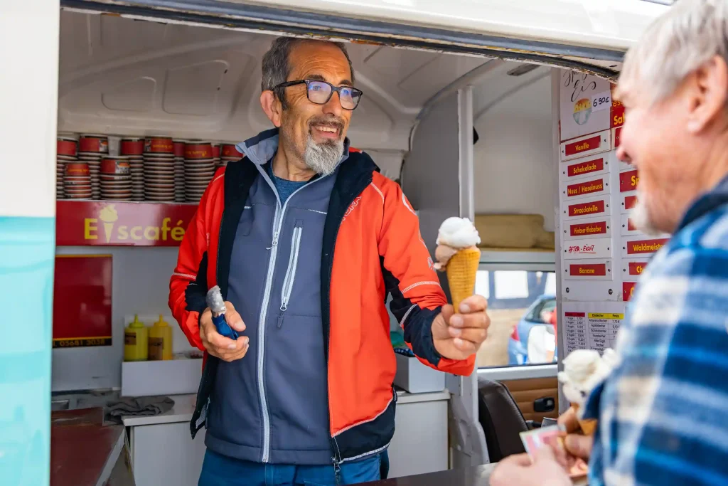 Ein freundlicher Mann reicht aus seinem Eiswagen eine Eistüte an einen Kunden.