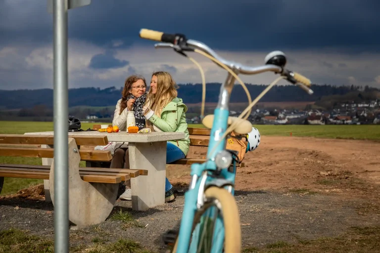 Zwei Frauen sitzen auf einer Parkbank in der Natur und genießen einen Snack, im Vordergrund steht ein Fahrrad. ©David Heise